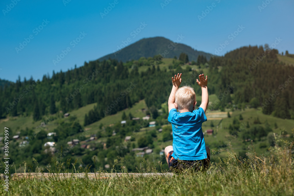 Little boy sits on mountains background with hands raised up. Back view Little boy sits on mountains background with hands raised up. Back view