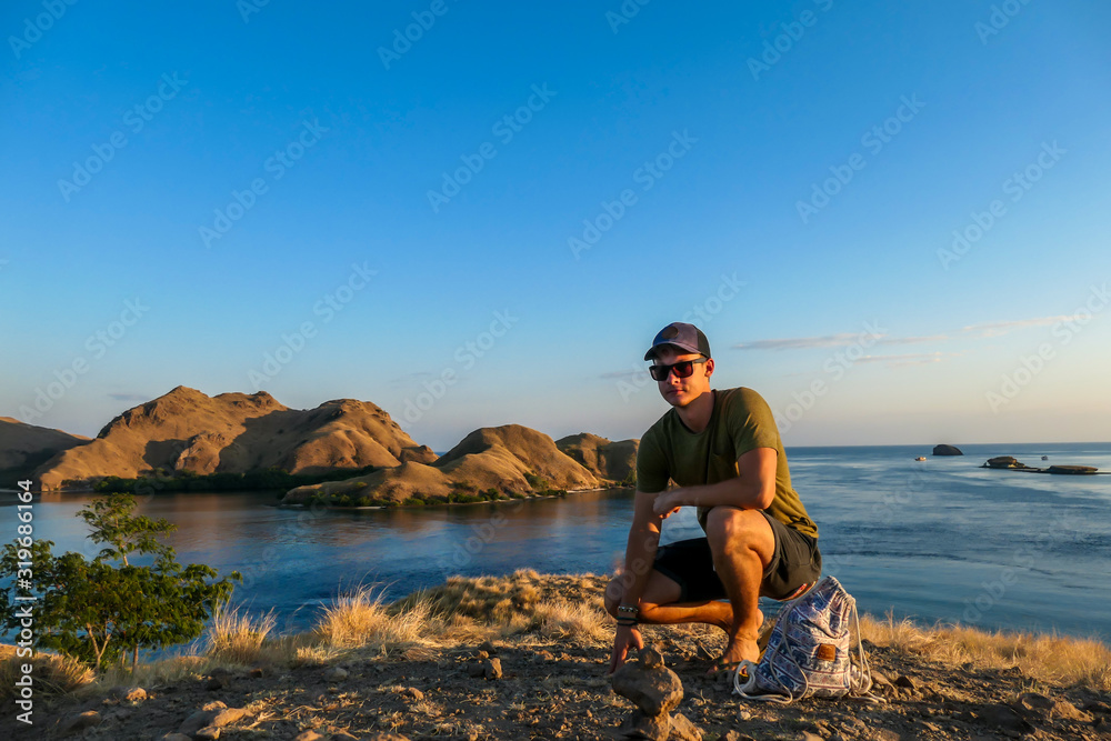 A man squatting on top of a small island, enjoying the morning sun over Komodo National Park, Flores, Indonesia. Golden hour over the islands and sea. Some boats anchored to the bay. New day beginning