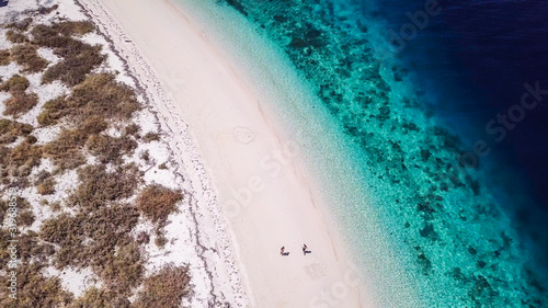 A drone shot of a couple playing on a pink sand beach on a small island near Maumere, Indonesia. Happy and careless moments. Waves gently washing the shore. Romance and love while travelling © Chris