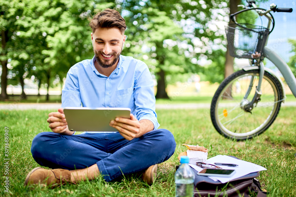 © baranq - Smiling young man sitting in park with tablet and bicycle during lunch break