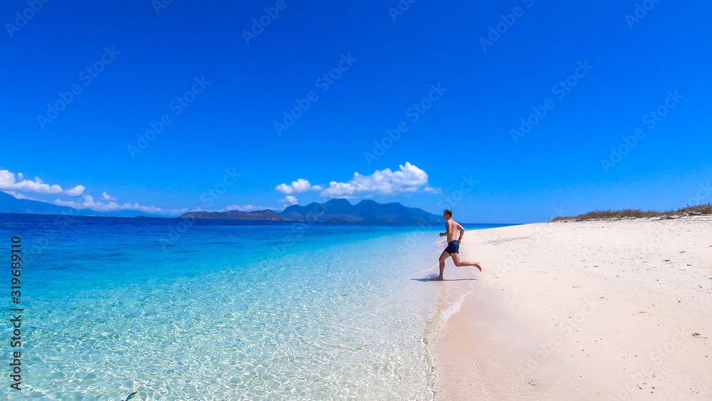 A man in swimsuit running into a crystal clear sea water on an island near Maumere, Indonesia. Clear, turquoise coloured water displaying coral reef. Happiness and fun while travelling. Hidden gem.