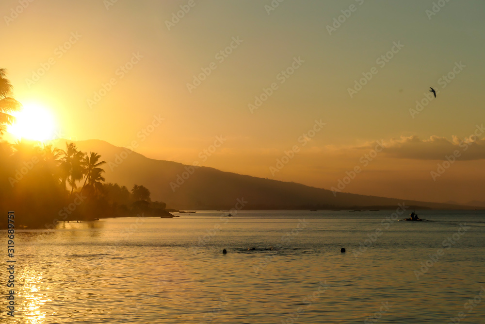 Obraz premium A sunset in Maumere, Flores, Indonesia. The sun sets behind the slope of the island. Serenity and calmness. There are few palm trees on the seashore. Few boats anchored to the bay.