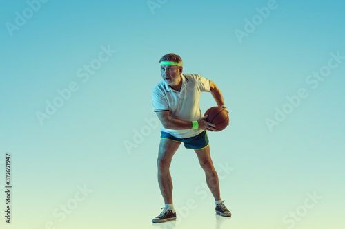 Senior man playing basketball on gradient background in neon light. Caucasian male model in great shape stays active, sportive. Concept of sport, activity, movement, wellbeing, healthy lifestyle.