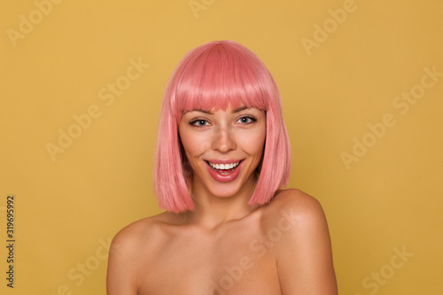 Indoor photo of beautiful young blue-eyed pink haired woman with bob haircut looking joyfully at camera and smiling widely while standing over mustard background