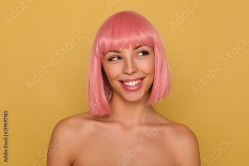 Portrait of young attractive pink haired female showing her perfect white teeth while smiling cheerfully, looking aside positively while standing over mustard background