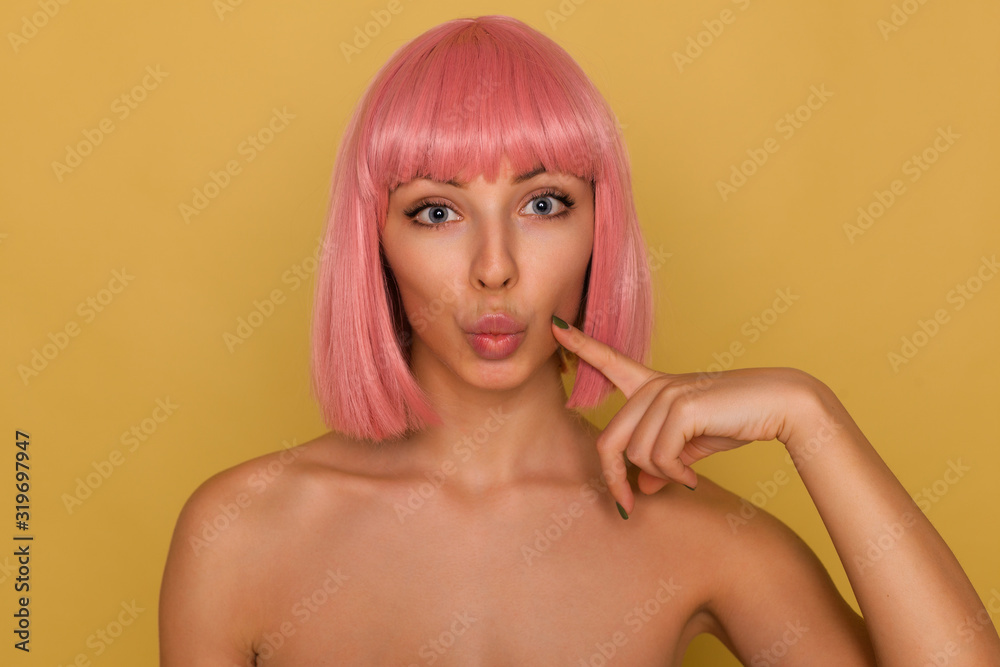 Obraz premium Close-up of young pretty blue-eyed pink haired woman with trendy bob haircut pouting her lips while looking at camera and keeping index finger on her cheek, isolated over mustard background