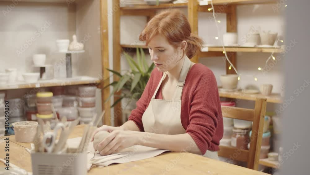 Waist-up arc shot of young Caucasian woman with red hair in ponytail, wearing apron, sitting at table in pottery workshop in front of supply storage racks, holding ball of clay and shaping it by hand Waist-up arc shot of young Caucasian woman with red hair in ponytail, wearing apron, sitting at table in pottery workshop in front of supply storage racks, holding ball of clay and shaping it by hand
