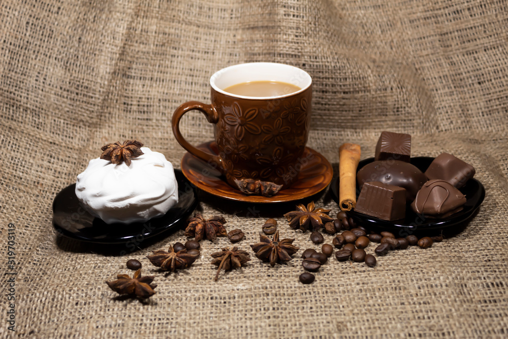 On the table is a cup of coffee with milk, a saucer with chocolates and marshmallows. Nearby are aniseles, coffee beans and cinnamon sticks. On the table is a cup of coffee with milk, a saucer with chocolates and marshmallows. Nearby are aniseles, coffee beans and cinnamon sticks.