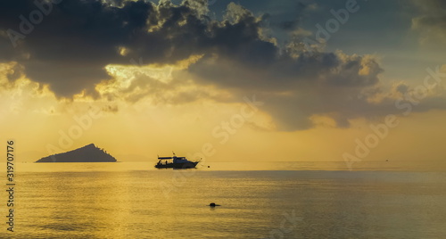 sea view panorama of a small boat floating in the sea with small island and sun rays with yellow sun light in cloudy sky background, sunrise at Ko Bulon Le island, Stun Province, southern of Thailand.