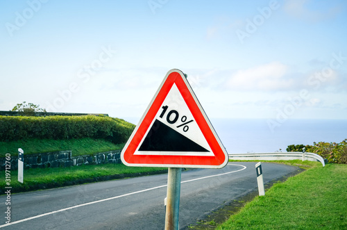 Red triangle road sign indicating a steep 10 percent downhill gradient in the road ahead. Empty road with crash barriers surrounded by green grass in the background. Traffic signs