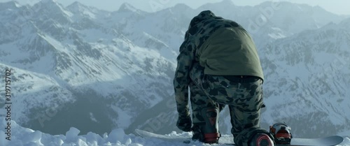 Back view of a professional snowboarder buckling into bindings on a mountain peak early in the morning, preparing for freeride snowboarding. 4K UHD, shot with anamorphic lens photo