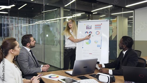 Attractive serious blond business worker explaining the graph using flip chart presentation for considerate high-skilled multiethnic business people in meeting room photo