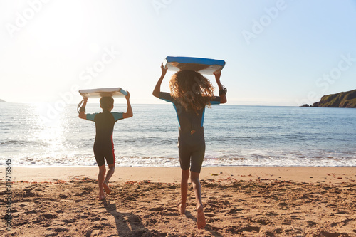Rear View Of Children In Wetsuits Carrying Bodyboards On Summer Beach Vacation Having Fun By Sea