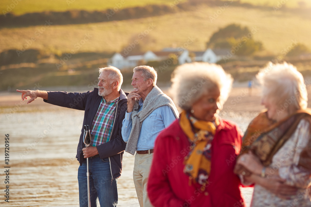 Group Of Smiling Senior Friends Walking Arm In Arm Along Shoreline Of Winter Beach