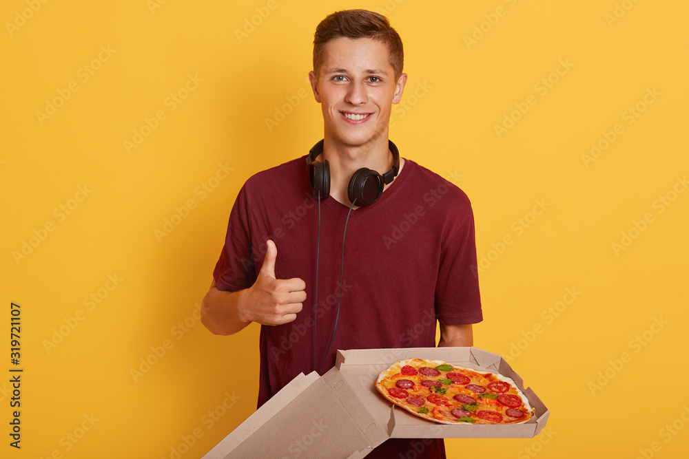 Indoor picture of young cheerful sweet boy standing isolated over yellow background in studio, holding opened cardboard box with pizza, making gesture, showing his approval, raising his thumb. Indoor picture of young cheerful sweet boy standing isolated over yellow background in studio, holding opened cardboard box with pizza, making gesture, showing his approval, raising his thumb.