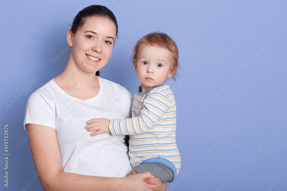 Studio shot of beautiful young mother standing and holding her baby boy in her arms in front of blue background, lady with dark hair and ponytail with cute infant, looking ar camera and smiling. Studio shot of beautiful young mother standing and holding her baby boy in her arms in front of blue background, lady with dark hair and ponytail with cute infant, looking ar camera and smiling.