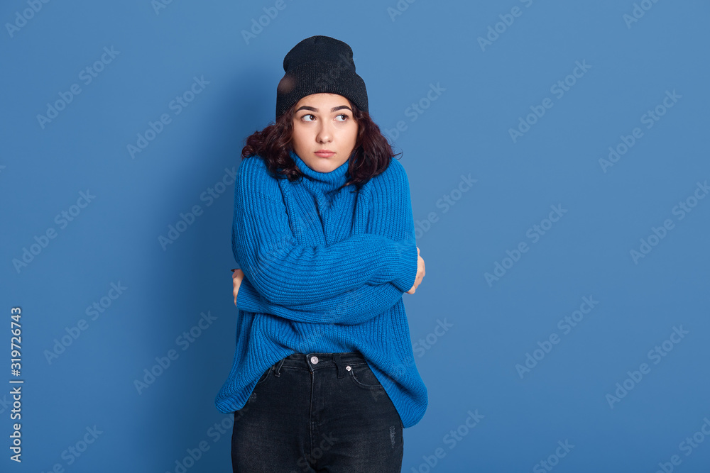Horizontal studio picture of emotional upset female standing isolated over blue background, looking aside, hugging herself, feeling cold, winter concept, wearing warm sweater, hat and trousers. Horizontal studio picture of emotional upset female standing isolated over blue background, looking aside, hugging herself, feeling cold, winter concept, wearing warm sweater, hat and trousers.