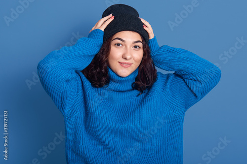 Horizontal picture of good looking beautiful female wearing dark blue and sweater, looking directly ar camera, putting hands on head, having curly black hair, being in high spirits. Winter concept. photo