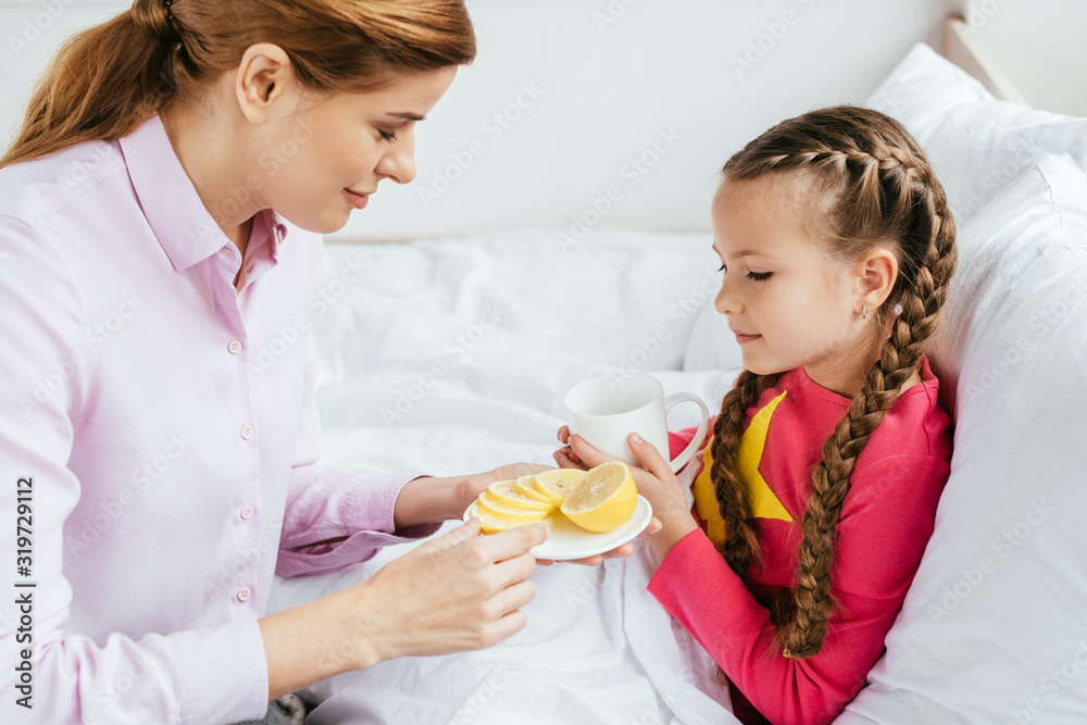 smiling mother giving lemon to ill daughter with cup of hot drink