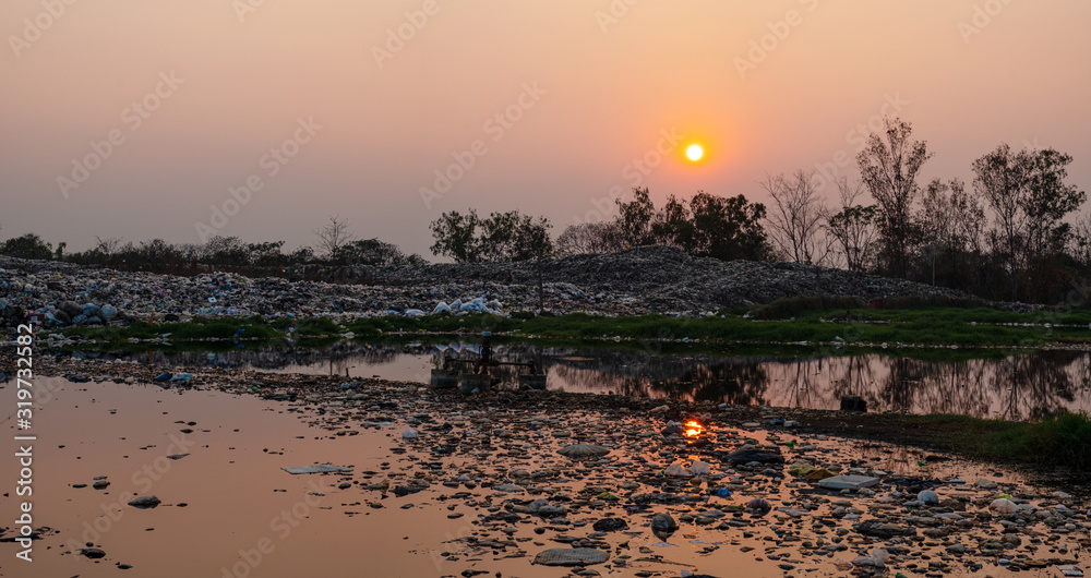 Naklejka premium Polluted water and mountain large garbage pile and pollution at the sun is setting in the background