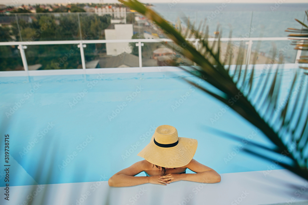 Obraz premium Stylish young woman in hat relaxing in pool under palm leaves and enjoying summer holiday. Summer vacation. Girl in sunhat on vacation in luxury tropical resort, swimming in pool on rooftop