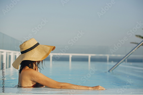 Stylish young woman in hat relaxing in pool and enjoying summer holiday. Summer vacation. Girl in wet sunhat on vacation in luxury tropical resort, swimming in pool on rooftop. Tranquil moment