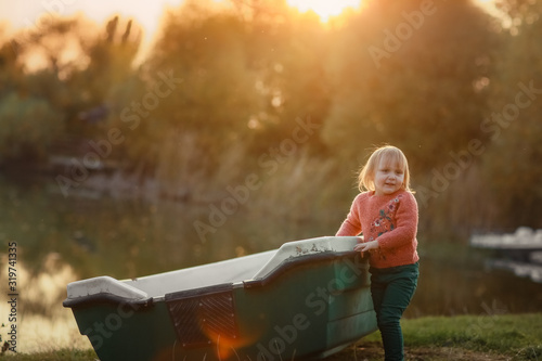 Child outdoors stands near a wooden boat at the lake.