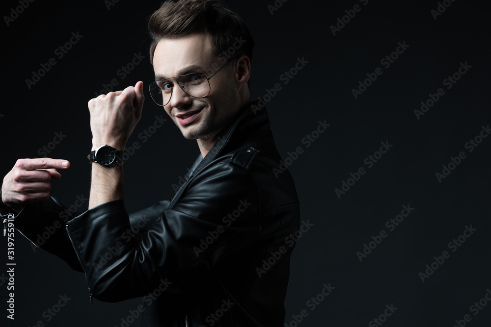 side view of smiling stylish brutal man in biker jacket pointing with finger at wristwatch isolated on black