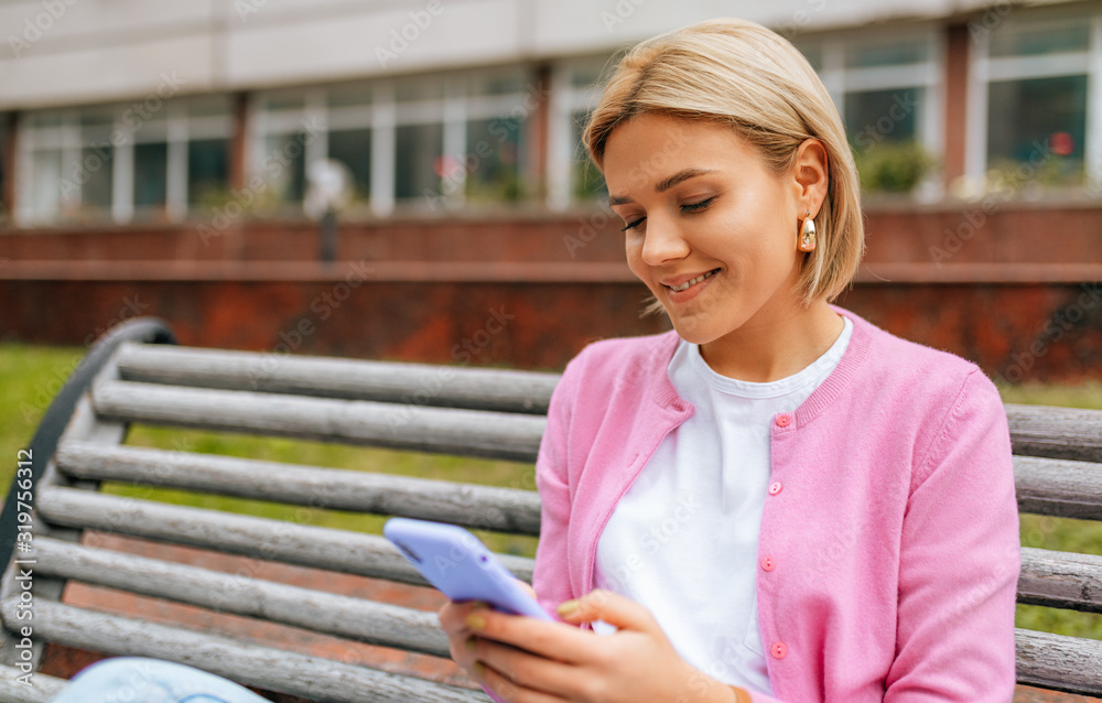 Smart blonde young woman wearing blue jeans, white t-shirt and pink jacket, smiling and sending messages on smart phone. Pretty female browsing on the web in the city street in spring time. Smart blonde young woman wearing blue jeans, white t-shirt and pink jacket, smiling and sending messages on smart phone. Pretty female browsing on the web in the city street in spring time.