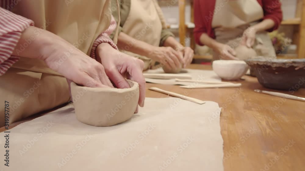 Close-up shot of hands of three unrecognizable women in aprons standing at table in pottery workshop and sculpting handmade earthenware bowls from raw clay