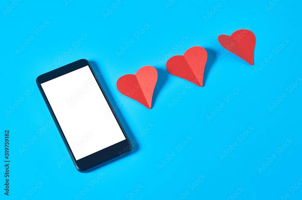 Black smartphone with isolated white screen for text, picture, photo and other graphics near row of three red paper hearts on blue table. Valentines day and love concept. Space for text. Close-up Black smartphone with isolated white screen for text, picture, photo and other graphics near row of three red paper hearts on blue table. Valentines day and love concept. Space for text. Close-up