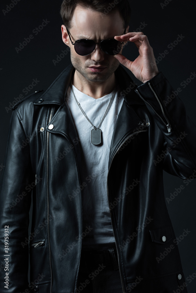 Obraz premium stylish brutal man posing in biker jacket and sunglasses isolated on black