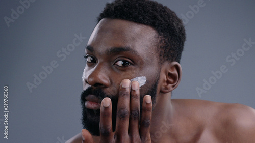 Close-up of handsome young sexy shirtless man applying moisturizing cream on face looking at camera. Nude portrait of attractive guy caring for his skin preparing for a day.