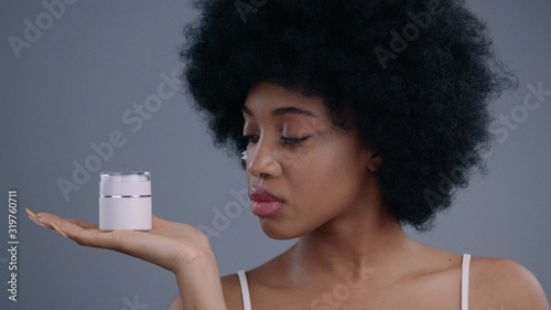 Portrait of gorgeous african american young woman with a cream on a cleansed tender face. Beautiful girl with curly hair demostrating skin care cream bottle looking at camera. photo