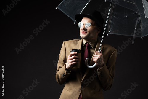 confused elegant man in beige suit, hat and eyeglasses holding umbrella and coffee to go isolated on black