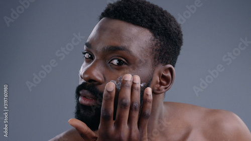 Portrait of attractive african young man using a moisturizing facial cream gently touching his face looking in the mirror. Close-up of nice-looking guy demostrating a jar of cream smiling at camera. © Fractal Pictures