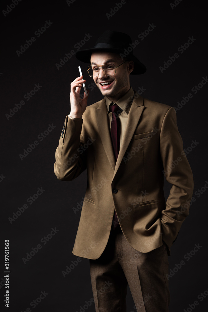Obraz premium smiling elegant man in beige suit, hat and eyeglasses talking on smartphone isolated on black