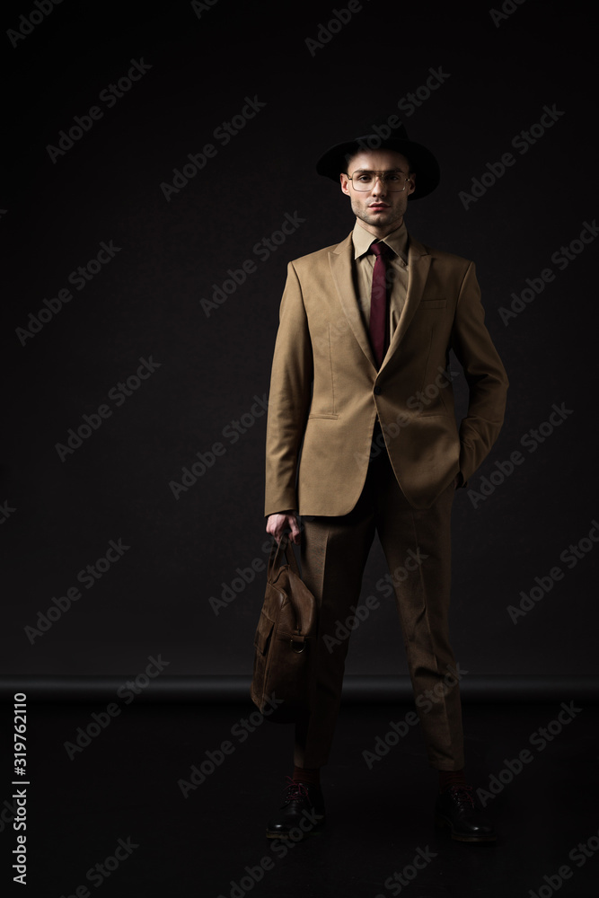 elegant man in beige suit, hat and eyeglasses holding brown leather bag with hand in pocket on black elegant man in beige suit, hat and eyeglasses holding brown leather bag with hand in pocket on black