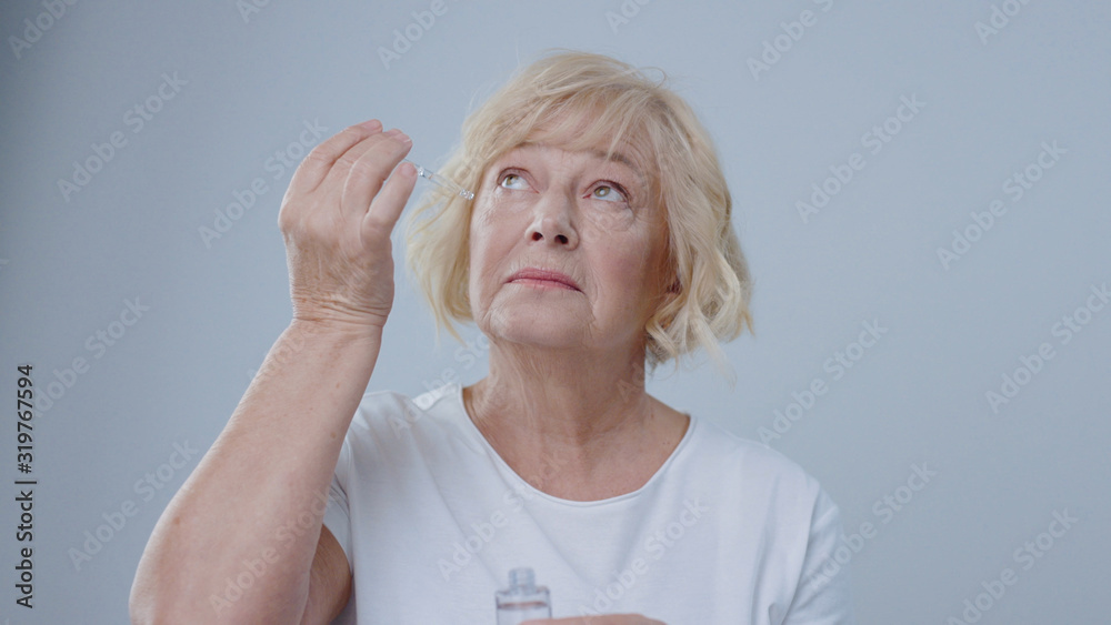 Blonde older woman putting moisturizer on her clean face pretending to cry isolated on grey background. Blonde older woman putting moisturizer on her clean face pretending to cry isolated on grey background.