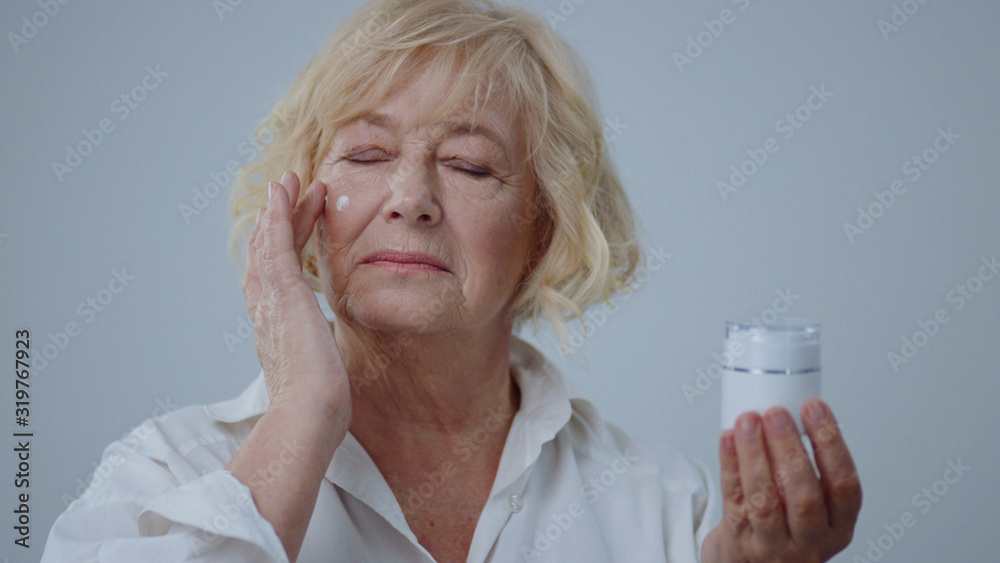 Portrait of cheerful older woman applying moisturizer to her face looking in the mirror. Wonderful senior model with blonde hair in white shirt holding cream isolated on grey background. Portrait of cheerful older woman applying moisturizer to her face looking in the mirror. Wonderful senior model with blonde hair in white shirt holding cream isolated on grey background.
