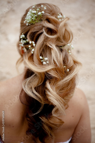 Blond woman with beautiful log hair on nude shoukders. The hairstyle of bride qith flowers , back view of salon hair do against wall. Care and beauty of hair