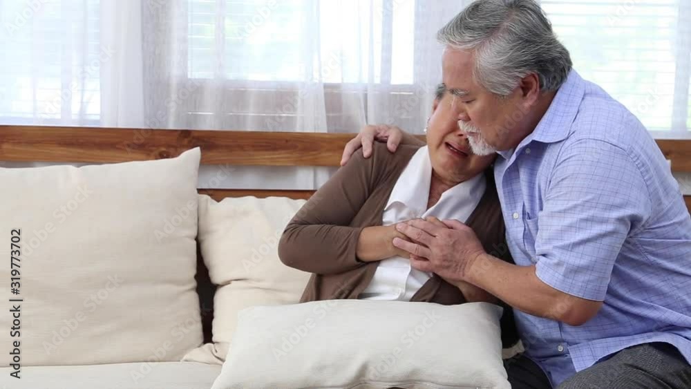 Asian senior woman with pain on heart on sofa in living room and take care by her husband, Health problem and feeling sick Asian senior woman with pain on heart on sofa in living room and take care by her husband, Health problem and feeling sick