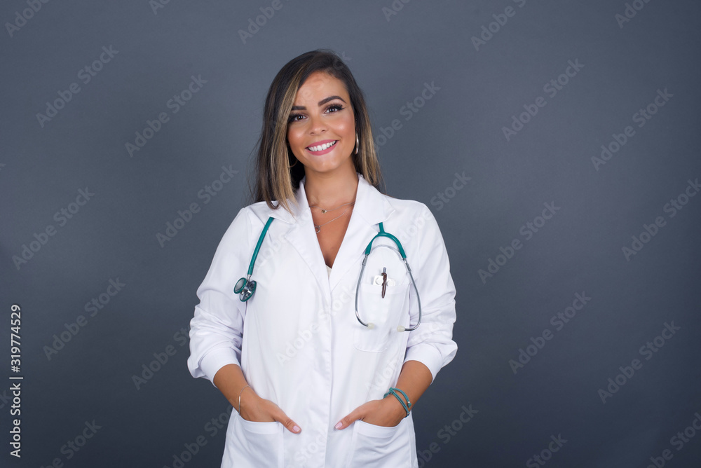 Confidence and business concept. Portrait of charming successful young doctor girl, smiling broadly with self-assured expression while holding hands in pockets standing against gray wall. Confidence and business concept. Portrait of charming successful young doctor girl, smiling broadly with self-assured expression while holding hands in pockets standing against gray wall.