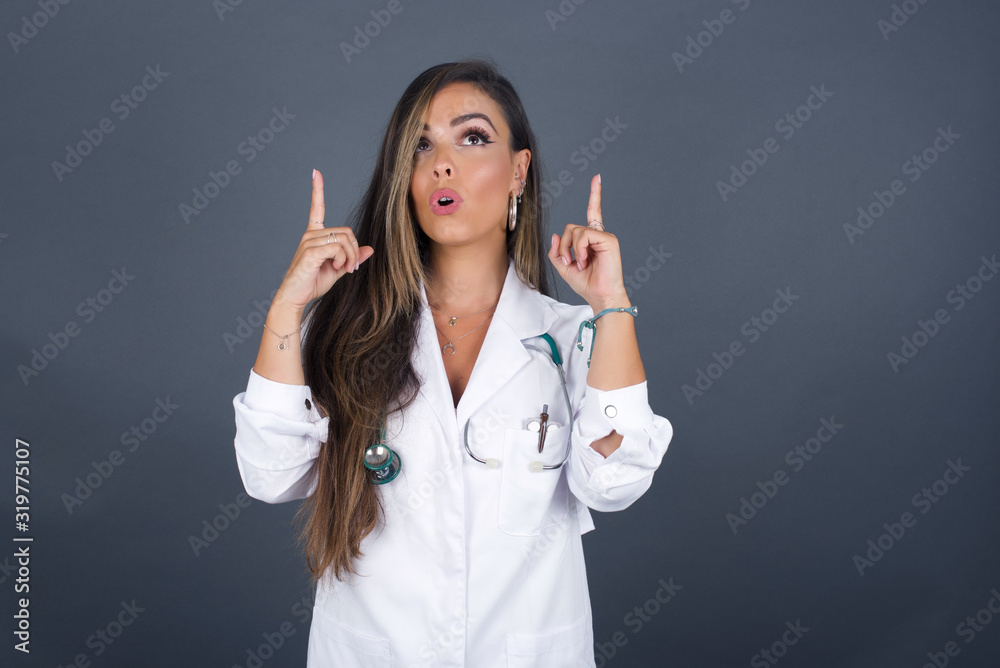 Portrait of beautiful doctor woman over isolated background being amazed and surprised looking and pointing up with fingers showing something strange. Portrait of beautiful doctor woman over isolated background being amazed and surprised looking and pointing up with fingers showing something strange.