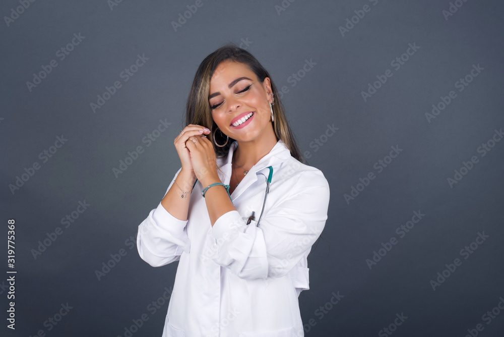 Dreamy young doctor woman with pleasant expression, closes eyes, keeps hands crossed near face, thinks about something pleasant, poses against gray background. Dreamy young doctor woman with pleasant expression, closes eyes, keeps hands crossed near face, thinks about something pleasant, poses against gray background.