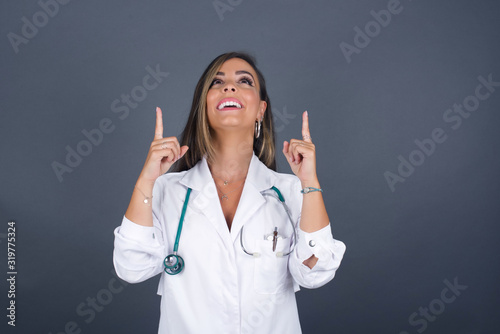 Charming carefree doctor woman with positive expression  points up with both index fingers  dressed medical uniform  has broad interested smile  isolated over gray background. Look there  please.