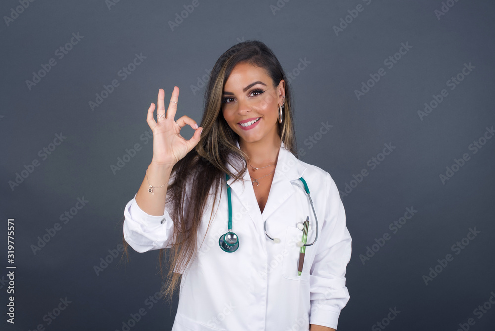 Glad attractive doctor woman shows ok sign with hand as expresses approval, has cheerful expression being optimistic. Standing against gray wall. Glad attractive doctor woman shows ok sign with hand as expresses approval, has cheerful expression being optimistic. Standing against gray wall.