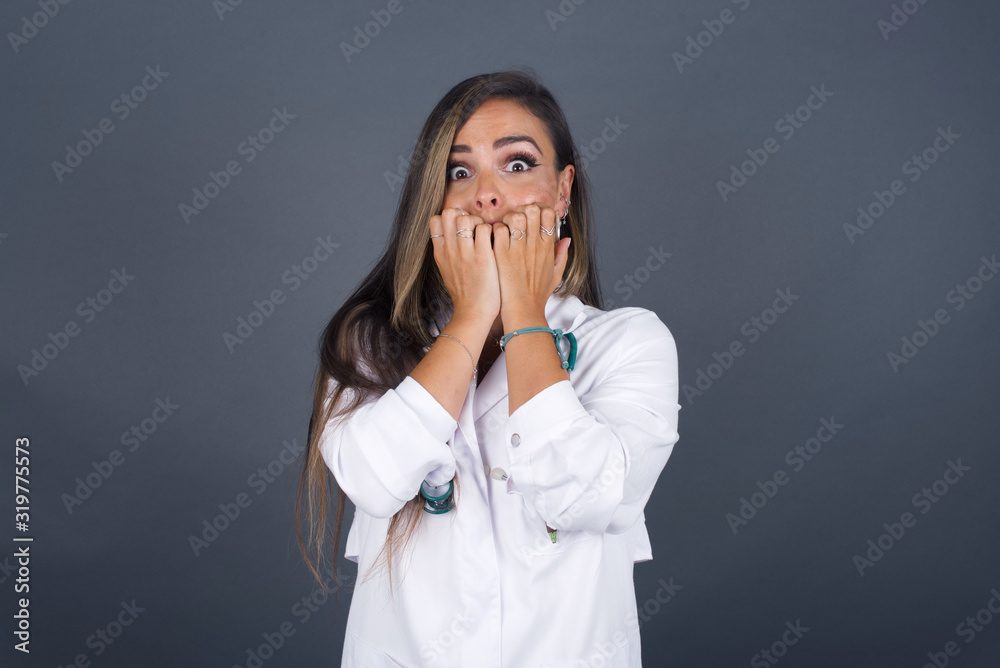 Anxiety. beautiful young caucasian doctor woman covering her mouth with her hands and standing indoors. Scared from something or someone bitting nails. Anxiety. beautiful young caucasian doctor woman covering her mouth with her hands and standing indoors. Scared from something or someone bitting nails.
