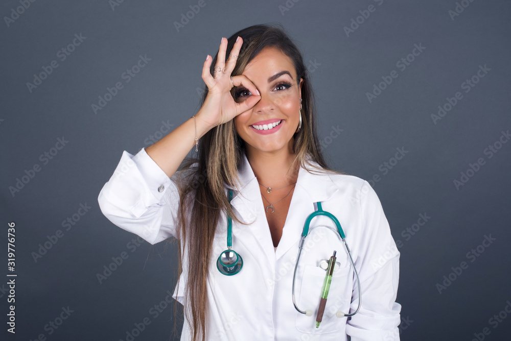 Beautiful young doctor female with happy face smiling doing ok sign with hand on eye looking through finger. Wearing medical uniform standing against gray wall. Beautiful young doctor female with happy face smiling doing ok sign with hand on eye looking through finger. Wearing medical uniform standing against gray wall.