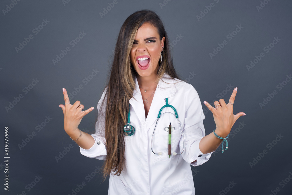 Born to rock this world. Joyful doctor woman in medical uniform screaming out loud and showing with raised arms horns or rock gesture, expressing excitement of being on concert of band. Born to rock this world. Joyful doctor woman in medical uniform screaming out loud and showing with raised arms horns or rock gesture, expressing excitement of being on concert of band.